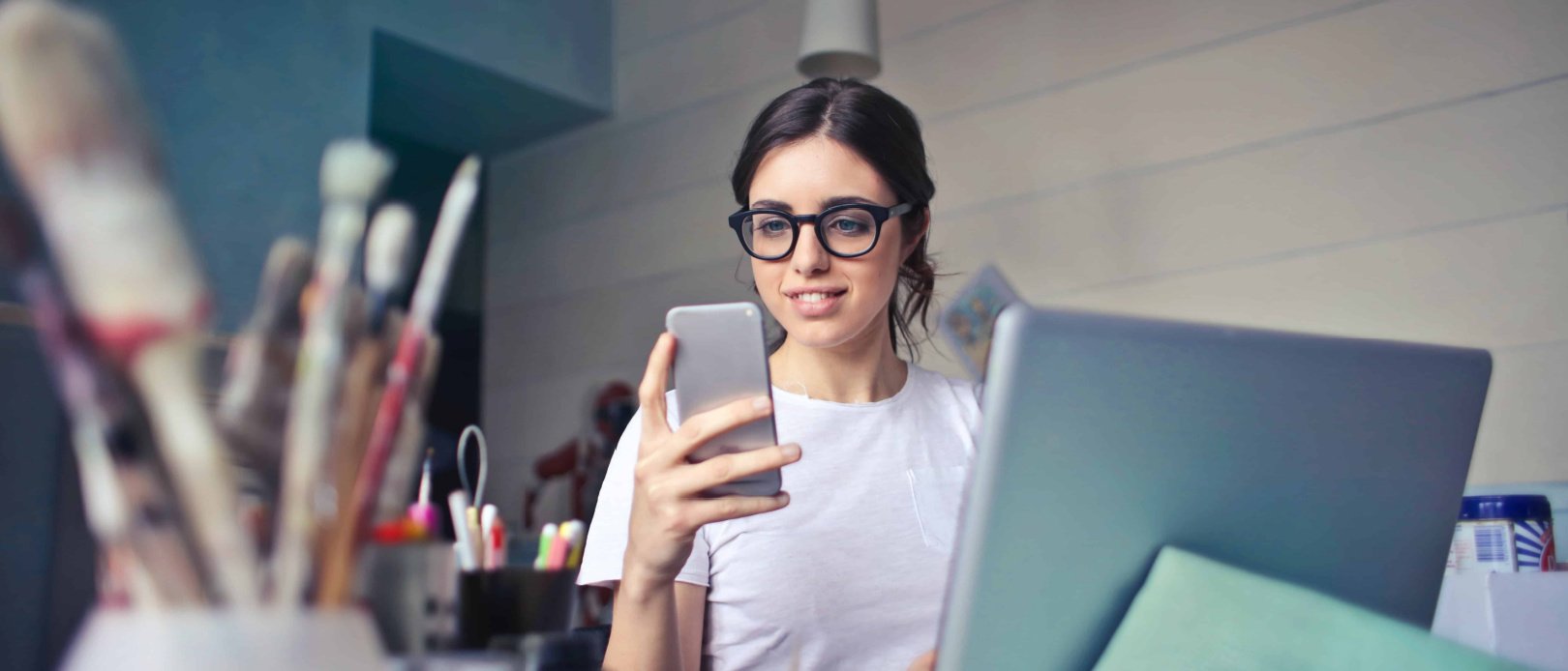 woman in glasses sitting at messy desk looking at mobile phone comparing